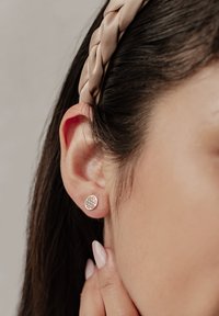 Orecchini tondi in argento con complessi motivi geometrici intagliati, indossati da una persona, insieme a una fascia intrecciata beige e smalto per unghie neutro.