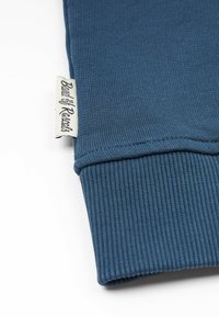 Ribbade blå mjuka tyget med uppvikt mudd och en liten vävd etikett med texten "Built by Renalt", som visar tydliga sömnadsdetaljer.