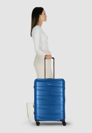 Donna con top bianco e pantaloni beige che tiene il manico di una valigia blu con ruote, in profilo su uno sfondo grigio.