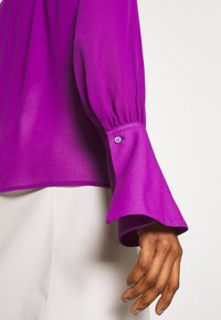 Lila, durchsichtige Bluse mit langen, gerafften Ärmeln und Knopfdetails an den Bündchen. Weicher Stoff und fließendes Design.