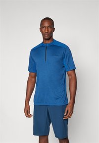 Camiseta atlética de manga curta em tecido azul mesclado com colarinho com zíper e textura leve que absorve a umidade. Combinada com calções azuis.