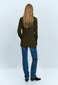 Blazer sur mesure vert olive au design épuré, associé à un jean en denim bleu taille haute et des bottines noires. Texture lisse, silhouette ajustée.
