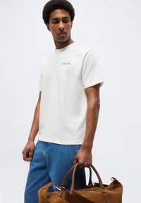 Άνδρας που φοράει λευκό μπλουζάκι με το κείμενο "JUICE IT UP", μπλε τζιν, κρατώντας καφέ δερμάτινη τσάντα χειρός από τις λαβές της.