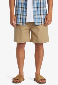Khaki šortky s hladkou strukturou, v kombinaci s modrou kostkovanou košilí. Hnědé klouzavé sandály z měkkého materiálu s minimalistickým designem.