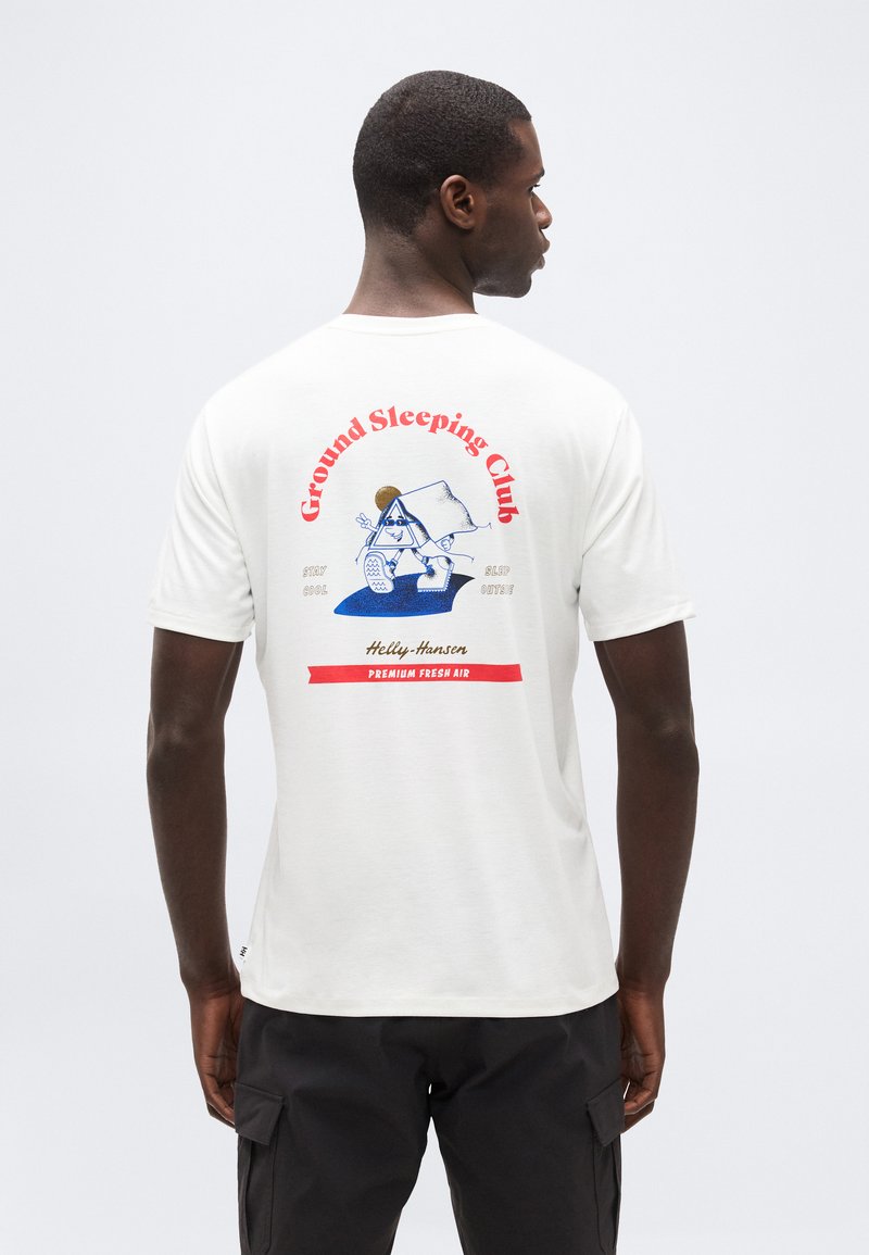 Άνδρας που φοράει λευκό μπλουζάκι με πολύχρωμο σχέδιο και κόκκινο κείμενο "Ground Sleeping Club" και μαύρο παντελόνι, στέκεται με την πλάτη προς το μέρος μας μπροστά σε απλό φόντο.