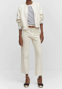 Giacca bomber bianca con texture, camicia bianca e blu a righe, pantaloni corti beige, cintura marrone e sandali neri flat con accenti dorati.
