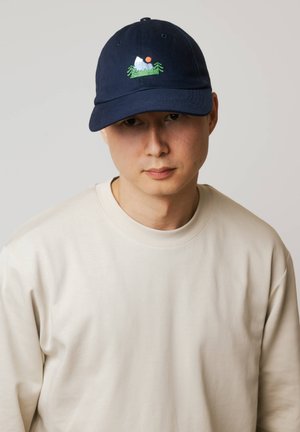 Mężczyzna w granatowej czapce z haftem przedstawiającym górę, drzewa i słońce oraz w beżowej bluzie z okrągłym dekoltem na jasnym, jednolitym tle.