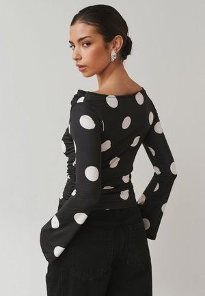 Mujer con el pelo oscuro recogido en un moño, lleva pendientes plateados con forma de concha y una camiseta negra de manga larga con grandes lunares blancos, manos en las caderas.