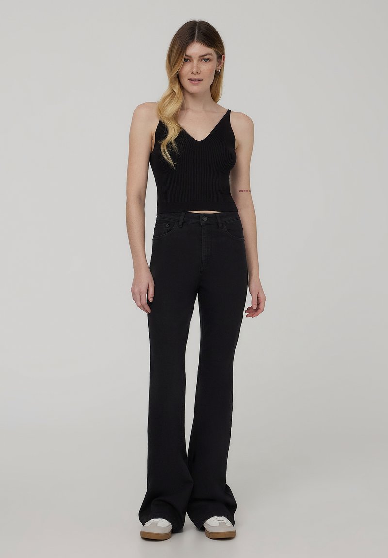 Top corto nero a costine con scollatura a V e design aderente, abbinato a jeans neri a zampa. Il modello indossa anche sneakers bianche con suola color beige.