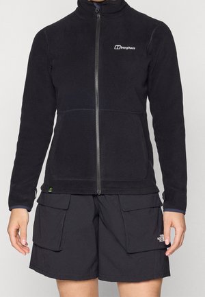 Person trägt eine schwarze Berghaus Fleecejacke mit Reißverschluss und schwarze The North Face Cargo-Shorts mit großen Fronttaschen.