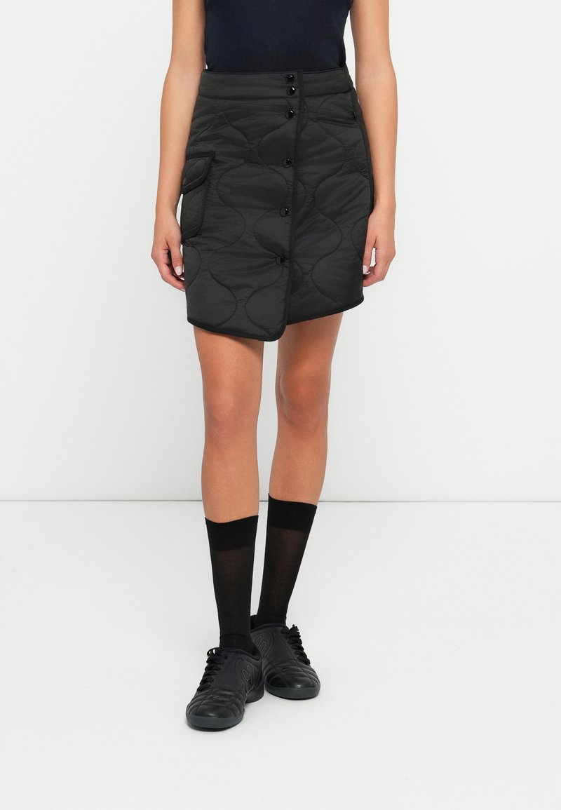 Jupe matelassée noire avec un devant à boutons, dotée d'une poche et d'un ourlet courbé. Portée avec des chaussettes noires montantes jusqu'au genou et des baskets noires.