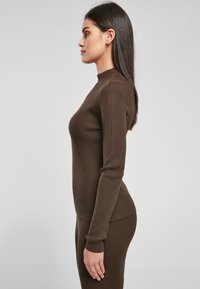 Brun ribbet rullekragegenser med lange ermer, tettsittende design og glatt tekstur, vist fra siden mot en lys grå bakgrunn.