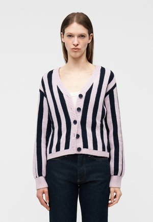 Kobieta ubrana w zapinany na guziki kardigan z pionowymi różowo-czarnymi paskami i długimi rękawami, w połączeniu z ciemnoniebieskimi dżinsami.