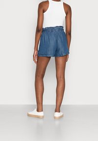 Denim-Shorts mit lockerem Schnitt, elastischem Bund und umgeschlagenen Säumen. Die Farbe ist ein mittleres Blau mit weicher Textur, kombiniert mit weißen Sneakern.