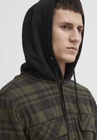 Grøn- og sortternet flannel skjorte med sort hætte, der har knaplukning og to store forlommer. Blød tekstur.