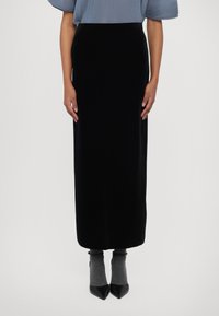 Schwarzer Samt-Maxikilt mit enganliegender Silhouette, kombiniert mit grauen gerippten Socken und spitzen schwarzen Absätzen. Glatte Textur und elegantes Design.