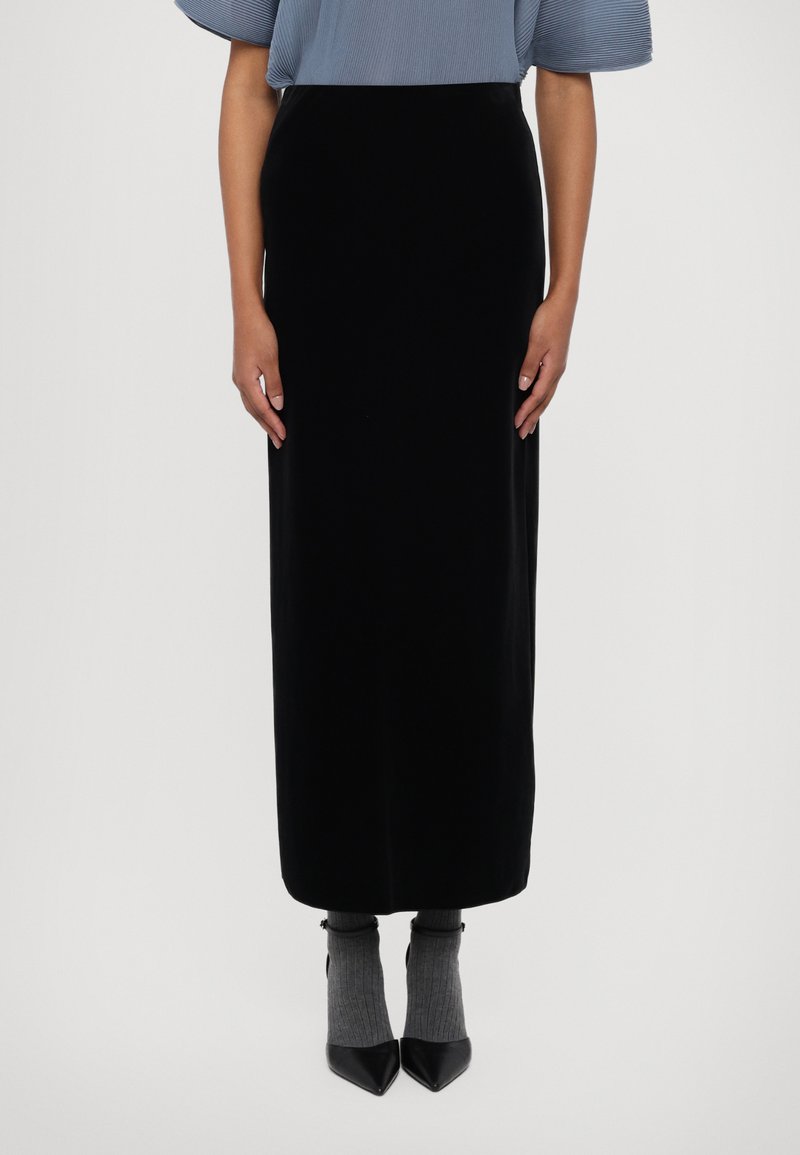 Schwarzer Samt-Maxikilt mit enganliegender Silhouette, kombiniert mit grauen gerippten Socken und spitzen schwarzen Absätzen. Glatte Textur und elegantes Design.