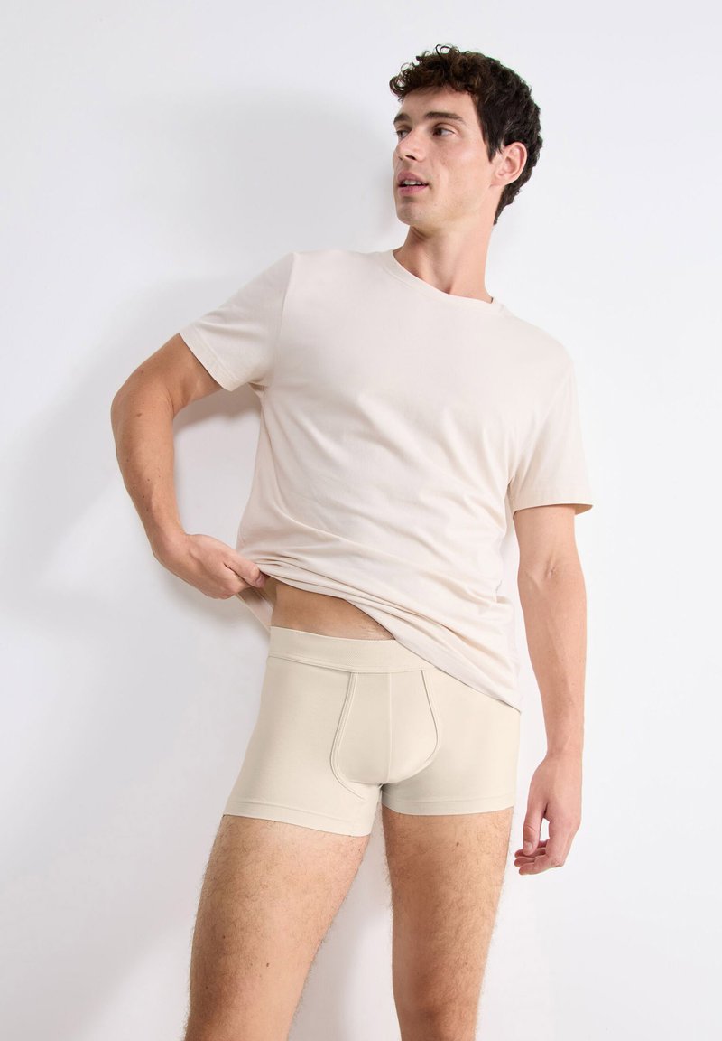 Boxer-briefs beiges en tissu doux, avec une coupe ajustée et une ceinture élastique, associés à une chemise à manches courtes beiges assortie.