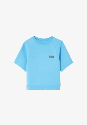 Garçon portant un sweat-shirt bleu clair avec le logo "American Vintage", une chemise à manches longues beige en dessous, et un pantalon gris clair, debout en position neutre.