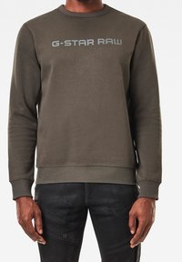 Sudadera verde oliva con cuello redondo, puños y dobladillo acanalados, con el logo "G-STAR RAW" en gris claro en el pecho. Textura suave.