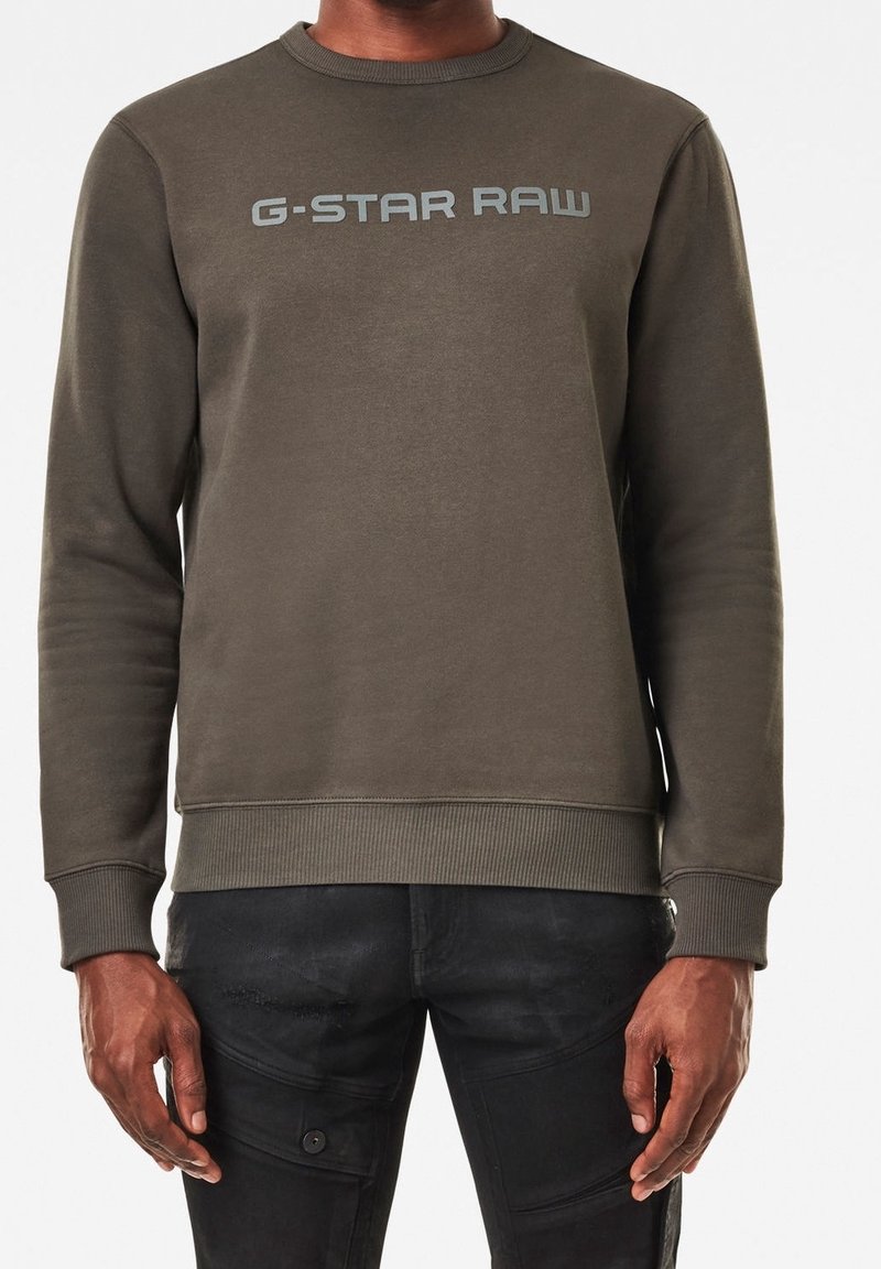 Sudadera verde oliva con cuello redondo, puños y dobladillo acanalados, con el logo "G-STAR RAW" en gris claro en el pecho. Textura suave.
