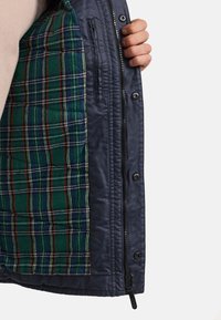 Veste marine avec une doublure à carreaux verte, bleue et rouge ; possède des boutons-pression et une poche zippée sur le côté à l'intérieur. Texture lisse.