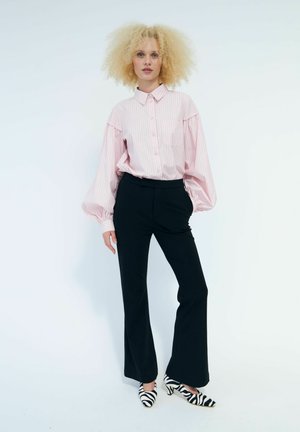 Donna con capelli biondi e ricci che indossa una camicetta rosa a righe, pantaloni neri a gamba larga e tacchi con stampa zebrata bianco e nero, in piedi su sfondo bianco.