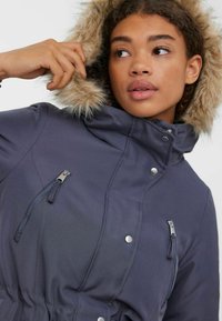 Mornarica parka z umetnim krznom na kapuci, zadrgo in gumbnimi zapirali, elastičnim pasom ter dvema sprednjima žepekoma z zadrgo. Gladko, teksturirano blago.