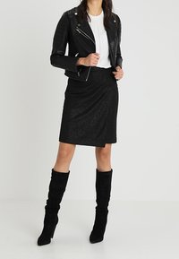 Zwarte leren bikerjack met gevoerde mouwen, witte geribde top, zwarte gestructureerde kokerrok en zwart suède laarzen tot de knie.