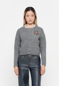 Cardigan grigio in maglia con scollatura tonda, dotato di cinque bottoni e ricamo di un orso sul lato sinistro del petto. Indossato sopra jeans in denim scuro.