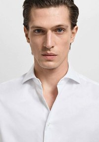 Camisa blanca de botón con cuello puntiagudo, textura suave y sin patrones visibles. El modelo tiene el cabello corto y peinado hacia atrás, con un fondo neutro.