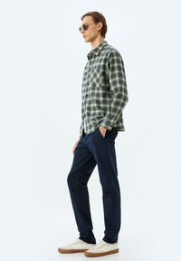 Grön och marinblå rutmönstrad skjorta med knapp, med en enda bröstficka, matchad med marinblå byxor och vita sneakers. Runda solglasögon bärs.