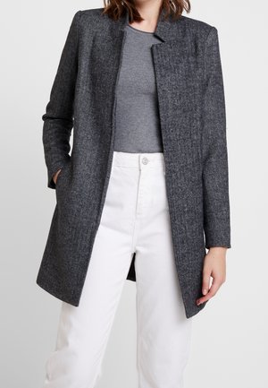 Kobieta w ciemnoszarym płaszczu w jodełkę, na wierzch założonym na szary prążkowany długi rękaw i białe spodnie z wysokim stanem, z ręką w kieszeni płaszcza.