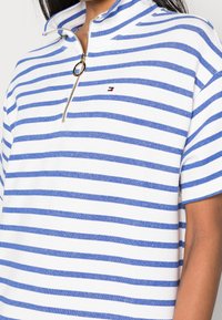 Polo w paski w kolorze białym i niebieskim, z zamkiem błyskawicznym przy dekolcie, krótkimi rękawami oraz małym haftowanym logo na piersi.