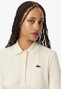 Camisa polo color crema con cuello acanalado, tapeta de tres botones y un pequeño logo bordado en el lado izquierdo del pecho. Textura de algodón suave.