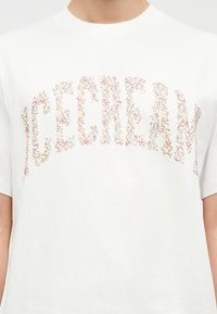 Λευκό μπλουζάκι με τη λέξη «ΕΛΕΥΘΕΡΙΑ» γραμμένη με πολύχρωμες κουφετίες στο στήθος, φορεμένο από ένα άτομο.