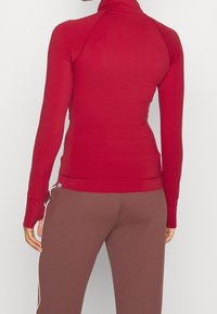 Rotes Langarmoberteil mit tailliertem Design, hohem Hals und glatter Textur, kombiniert mit braunen Hosen mit einem weißen Seitenstreifen.