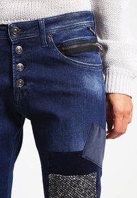 Jeans de mezclilla de color azul oscuro con un parche texturizado, un bolsillo con cremallera y múltiples botones plateados en la cintura.