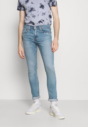 Mężczyzna w jasnoniebieskich, podwiniętych obcisłych dżinsach, białych trampkach i czarno-białej koszulce tie-dye, stojący na tle jasnej ściany.