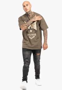 Olíva zöld grafikai póló merész "TAPOUT" felirattal, rövid ujjakkal, elnyűtt fekete farmerrel és fehér sneakers-szel párosítva.