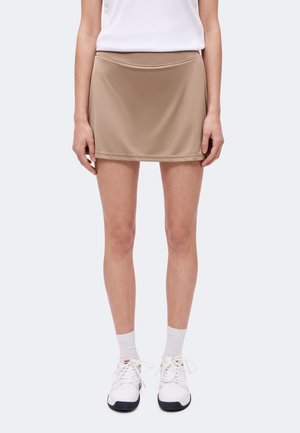 Persona que lleva una minifalda beige, camiseta blanca, calcetines blancos hasta la pantorrilla y zapatillas deportivas blancas, de pie frente a un fondo liso.