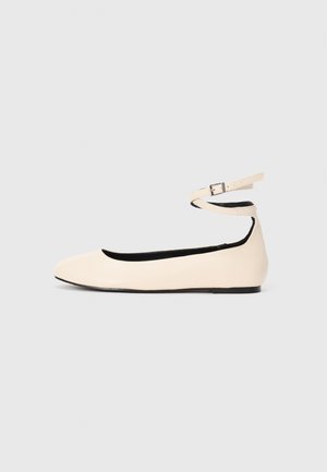 Vaalean beige tasapohjainen kenkä, jossa on pyöristetty kärki ja nilkkahihna, jossa on pieni hopeinen solki, vaalealla yksinkertaisella taustalla.