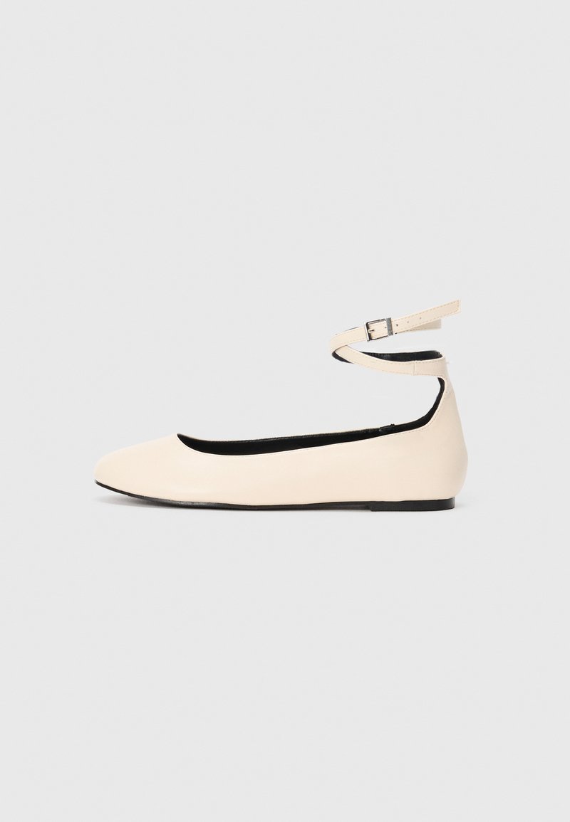 Botte plate beige avec un bout rond et une sangle au niveau de la cheville, présentant une petite boucle en argent, sur un fond clair.