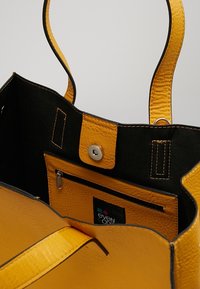 Gul læder tote taske med sort foring, der har en trykknapslukning, robuste håndtag og en indvendig lynlås-lomme til små genstande.