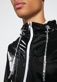Svart, glänsande, lättviktig jacka med dragsko i huvan, vit framfördragkedja och varumärkesdragband. Texturerad yta med en modern design.