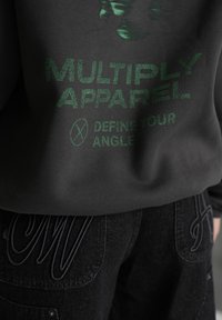 Sudadera negra con texto gráfico verde "MULTIPLY APPAREL" y "DEFINE YOUR ANGLE". Combinada con jeans oscuros con costuras en contraste.