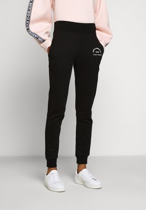 Persoon draagt zwarte joggingbroek met logo op dij, lichtroze sweater met tekst op mouwen en witte sneakers, staat met handen in zakken.