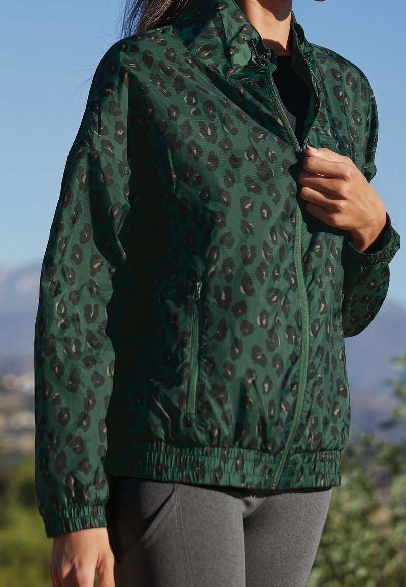 Femme portant une veste zippée verte à imprimé léopard et un pantalon gris, à l'extérieur avec des montagnes visibles en arrière-plan.