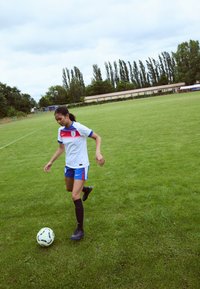 Jugador de fútbol con una camiseta blanca con acentos azules y rojos, pantalones cortos azules, calcetas negras y botines negros, caminando sobre hierba.