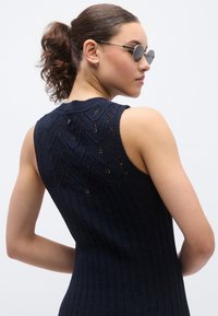 Μπλε σκούρο αμάνικο πλεκτό τοπ με ραβδωτή υφή και σχεδίαση σε σχήμα ζιγκ-ζαγκ στην πλάτη. Διαθέτει μικρό κούμπωμα με κουμπί στο λαιμό.